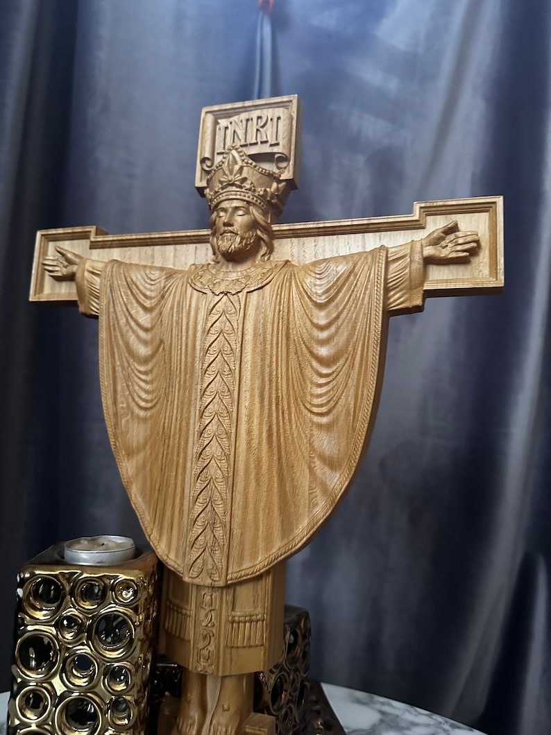 Może przedstawiać: Drewniana rzeźba krucyfiksu przedstawiająca ukoronowaną postać z wyciągniętymi ramionami, ubraną w szaty. Krzyż nosi napis "INRI". Rzeźba jest jasnobrązowa i spoczywa na dekoracyjnej podstawie.