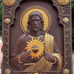 K&ouml;nnte beinhalten: Eine rechteckige Wandtafel aus dunkelbraunem Holz mit einem geschnitzten Relief des Heiligsten Herzens Jesu. Die Figur hat einen goldenen Heiligenschein und ein goldenes Herz mit Dornenkrone. Der Rahmen ist mit komplexen Mustern und Gold verziert.