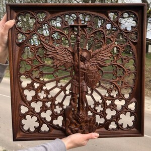 May include: A dark brown wooden wall panel featuring an intricate gothic rose window pattern. In the center, a carved figure of an angel with wings holds a sword and a shield inscribed with the Latin text "QUIS UT DEUS" while standing over a serpent.
