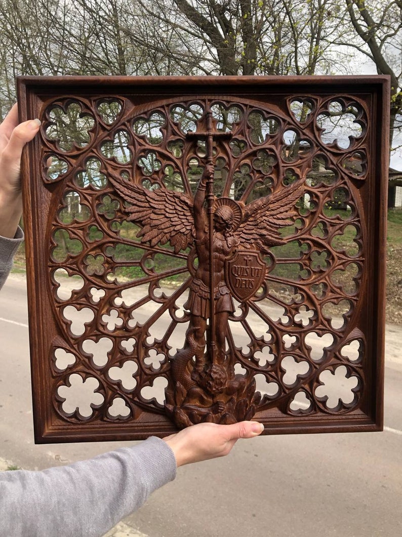 May include: A dark brown wooden wall panel featuring an intricate gothic rose window pattern. In the center, a carved figure of an angel with wings holds a sword and a shield inscribed with the Latin text "QUIS UT DEUS" while standing over a serpent.