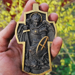 May include: A wooden cross-shaped religious pendant with a detailed relief carving of a figure in armor, holding a sword and shield. The cross has a gold-colored border and accents, with a halo around the figure's head. The pendant is held in a hand.