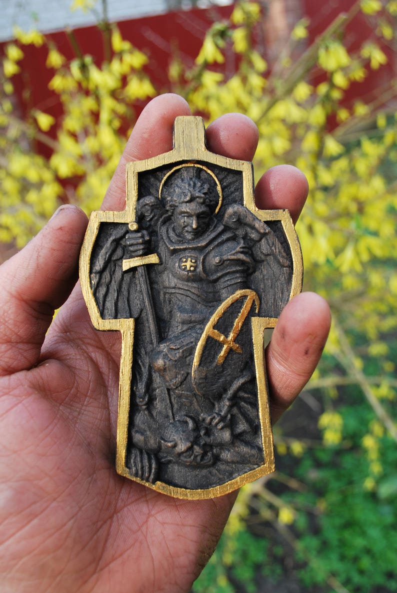 May include: A wooden cross-shaped religious pendant with a detailed relief carving of a figure in armor, holding a sword and shield. The cross has a gold-colored border and accents, with a halo around the figure's head. The pendant is held in a hand.