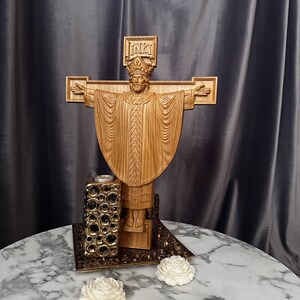 Może przedstawiać: Drewniana rzeźba religijna przedstawiająca Jezusa na krzyżu z wyciągniętymi ramionami. Rzeźba jest misternie wyrzeźbiona i stoi na dekoracyjnej podstawie. Obecny jest r&oacute;wnież świecznik i dwie białe dekoracje w kształcie r&oacute;ży.