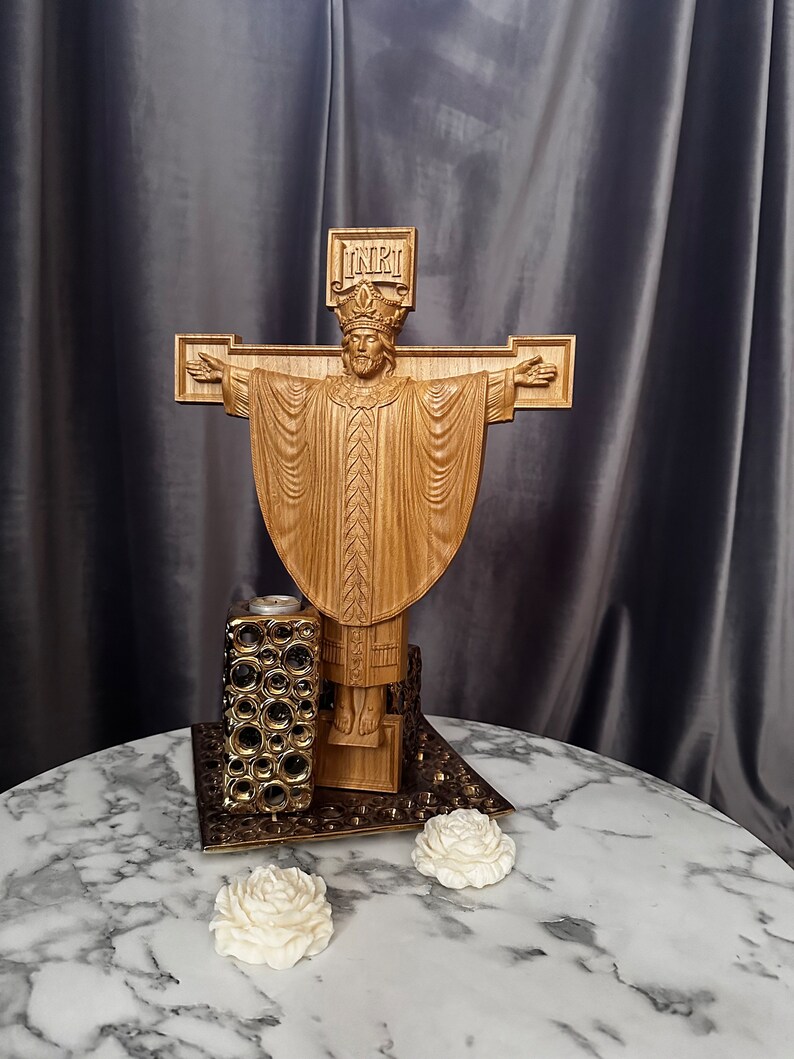 Może przedstawiać: Drewniana rzeźba religijna przedstawiająca Jezusa na krzyżu z wyciągniętymi ramionami. Rzeźba jest misternie wyrzeźbiona i stoi na dekoracyjnej podstawie. Obecny jest r&oacute;wnież świecznik i dwie białe dekoracje w kształcie r&oacute;ży.