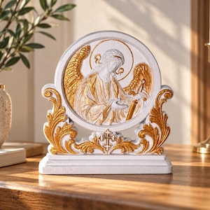 Op de afbeelding: Een wit en goudkleurig decoratief paneel met een reliëf van een engel met vleugels en een aureool. Het ronde centrale paneel is omlijst door sierlijk goudkleurig krulwerk en een klein kruis aan de basis, op een rechthoekig wit voetstuk.
