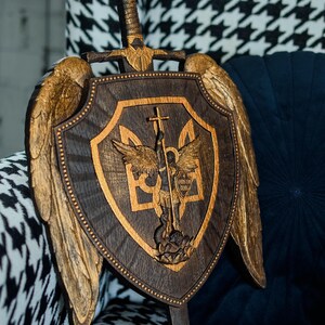 May include: A dark wood shield and sword wall plaque featuring gold-painted wings on the sides. The shield displays a central relief carving of a figure with wings holding a cross-topped sword over a trident symbol, with the Latin text Quis ut Deus.