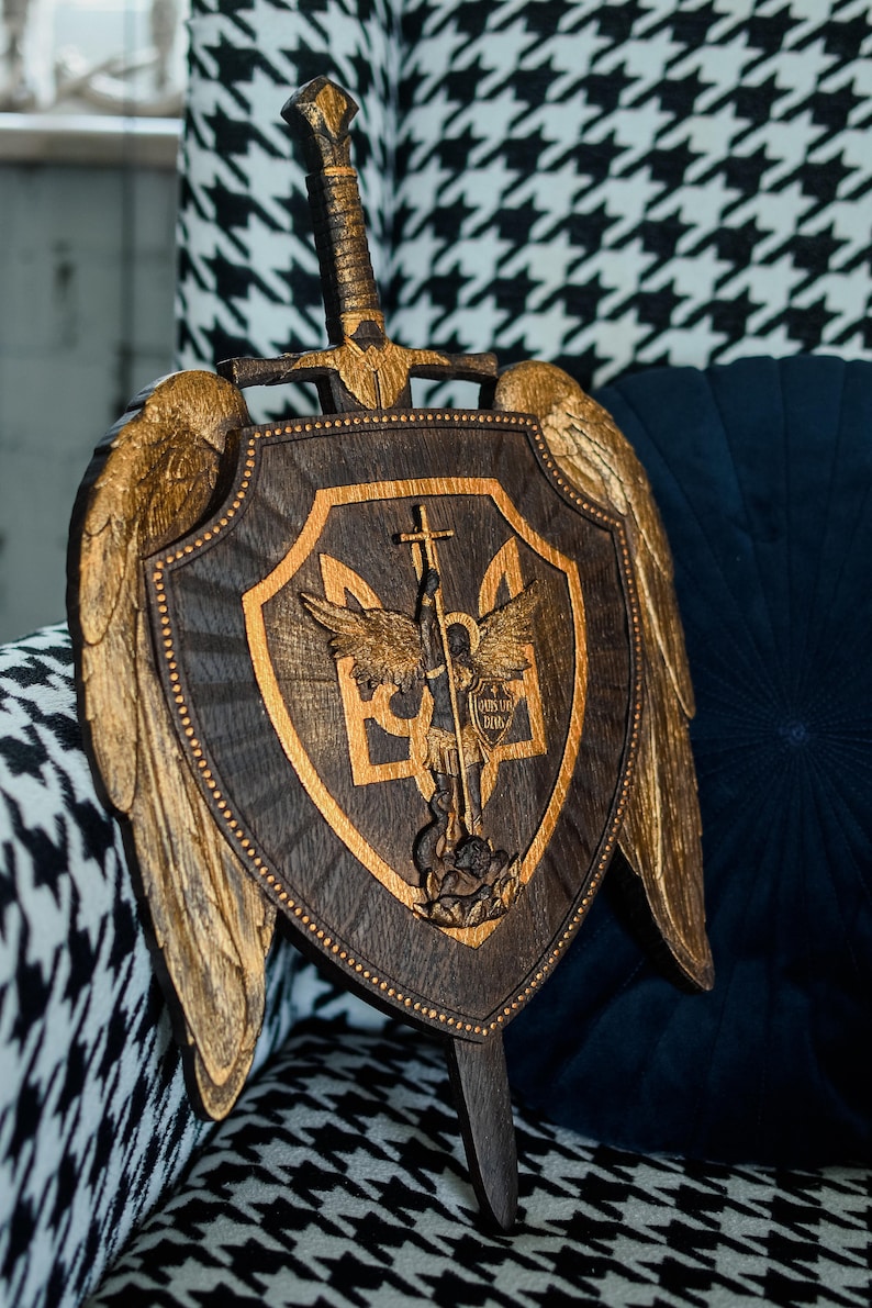 May include: A dark wood shield and sword wall plaque featuring gold-painted wings on the sides. The shield displays a central relief carving of a figure with wings holding a cross-topped sword over a trident symbol, with the Latin text Quis ut Deus.