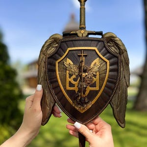 May include: A dark brown wooden shield plaque featuring a central gold-toned relief of Saint Michael the Archangel with wings, a sword, and the Ukrainian trident symbol. The shield is adorned with large gold-toned wings and a sword handle at the top.