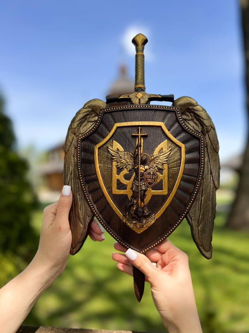 May include: A dark brown wooden shield plaque featuring a central gold-toned relief of Saint Michael the Archangel with wings, a sword, and the Ukrainian trident symbol. The shield is adorned with large gold-toned wings and a sword handle at the top.