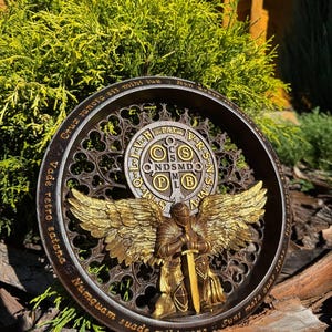 Puede incluir: Escultura en relieve circular detallada que presenta un ángel dorado con alas extendidas y una espada. El ángel está centrado dentro de un marco decorativo con inscripciones latinas. La escultura tiene una combinación de colores bronce y oro.