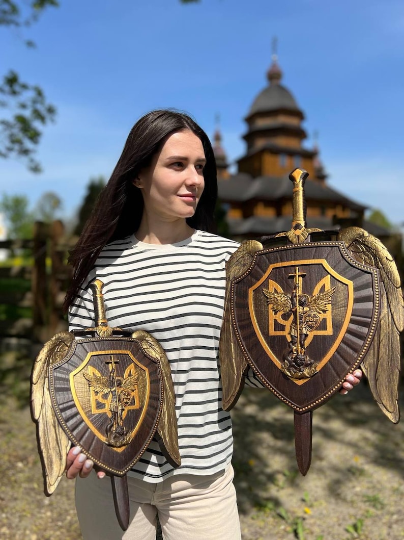 May include: Two dark brown wooden shields featuring gold-toned carved wings and a central sword. Each shield displays a gold relief of a winged figure standing over a serpent, set against the Ukrainian trident symbol on a dark wood background.