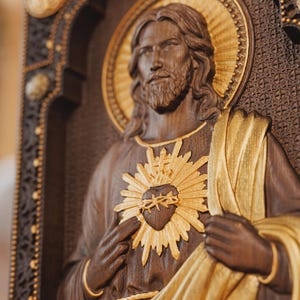 K&ouml;nnte beinhalten: Eine Wandtafel aus dunklem Holz mit einem geschnitzten Relief von Jesus Christus, einem goldenen Heiligenschein und einem goldenen Herz-Jesu-Emblem. Die Figur hat langes Haar und einen Bart und tr&auml;gt ein drapiertes Gewand mit Goldakzenten.