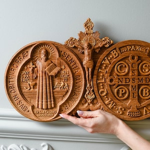 May include: A wooden wall plaque with three carved religious medallions. The center medallion features a crucifix with a figure of Jesus Christ. The medallions on either side feature the Saint Benedict Medal and the Miraculous Medal. The Saint Benedict Medal features a depiction of Saint Benedict holding a book and a cross. The Miraculous Medal features the Virgin Mary with a cross and the letters 'M', 'A', 'R', 'I', 'A' and 'S'.