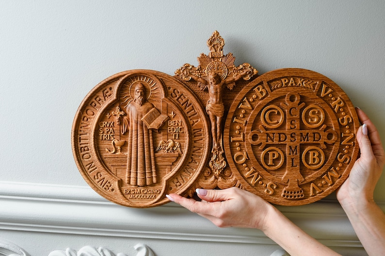 May include: A wooden wall plaque with three carved religious medallions. The center medallion features a crucifix with a figure of Jesus Christ. The medallions on either side feature the Saint Benedict Medal and the Miraculous Medal. The Saint Benedict Medal features a depiction of Saint Benedict holding a book and a cross. The Miraculous Medal features the Virgin Mary with a cross and the letters 'M', 'A', 'R', 'I', 'A' and 'S'.