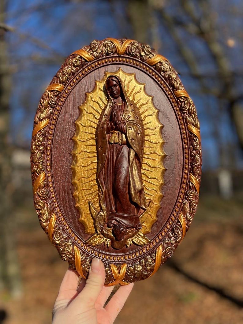 May include: An oval-shaped religious wall hanging featuring a carved figure of the Virgin Mary. The figure is depicted in a brown robe with a gold halo, set against a gold background. The frame is brown with gold accents.