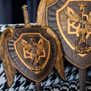 May include: Two dark brown wooden shields featuring gold-toned carved wings and a central relief of an angel with a sword and shield. The shield design includes the Ukrainian trident symbol and the Latin text "QUIS UT DEUS" on the angel's shield.