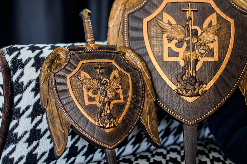 May include: Two dark brown wooden shields featuring gold-toned carved wings and a central relief of an angel with a sword and shield. The shield design includes the Ukrainian trident symbol and the Latin text "QUIS UT DEUS" on the angel's shield.