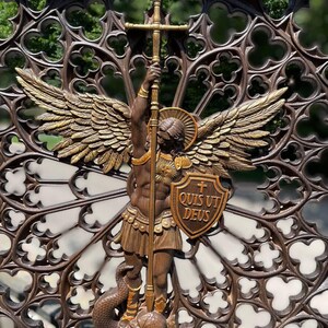 May include: A brown and gold-toned wall plaque featuring a winged figure holding a tall cross-topped staff and a shield inscribed with the Latin text "QUIS UT DEUS". The figure stands over a serpent-like form, set against a dark, intricate gothic-style circular lattice background with repeating quatrefoil cutouts.