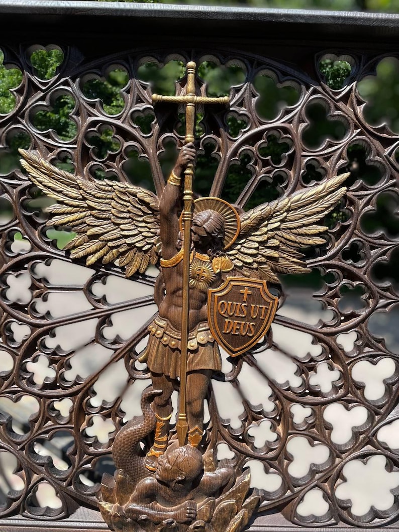 May include: A brown and gold-toned wall plaque featuring a winged figure holding a tall cross-topped staff and a shield inscribed with the Latin text "QUIS UT DEUS". The figure stands over a serpent-like form, set against a dark, intricate gothic-style circular lattice background with repeating quatrefoil cutouts.