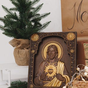 K&ouml;nnte beinhalten: Eine rechteckige Wandtafel aus Holz mit einem Reliefschnitzwerk des Heiligsten Herzens Jesu. Die Figur ist mit langem Haar, Bart und einem goldenen Heiligenschein dargestellt und h&auml;lt ein flammendes Herz. Der Rahmen hat detaillierte Schnitzereien.