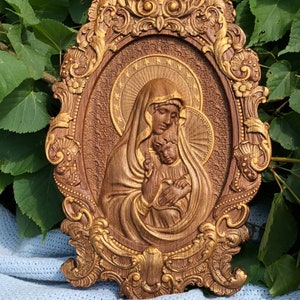 May include: An oval-shaped wooden wall plaque featuring a relief carving of a figure holding a child. The piece is framed by an ornate, decorative border with gold-toned highlights and a textured background featuring a halo with stars.