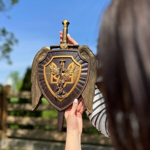 May include: A dark brown wooden wall plaque shaped like a shield with a sword handle extending from the top. It features gold-colored wings on the sides and a central gold emblem depicting an angel with a sword and the Ukrainian trident symbol.