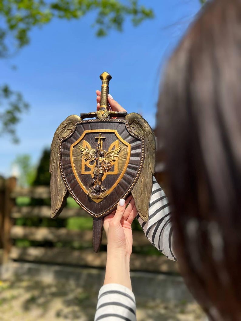 May include: A dark brown wooden wall plaque shaped like a shield with a sword handle extending from the top. It features gold-colored wings on the sides and a central gold emblem depicting an angel with a sword and the Ukrainian trident symbol.