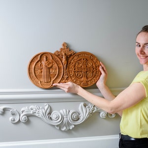 May include: A woman holds a wooden carving of Saint Benedict's Medal. The medal is a circular design with a cross in the center and the words "EGO VOX CLAMANS IN DESERTO" around the edge. The carving is a beautiful example of traditional craftsmanship.