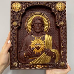 K&ouml;nnte beinhalten: Eine rechteckige Wandtafel aus Holz mit einem geschnitzten Relief des Heiligsten Herzens Jesu. Die Figur ist mit einem goldenen Heiligenschein und einem goldenen Herzsymbol dargestellt. Der Rahmen ist detailliert mit dunklem Holz und Gold.