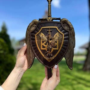 May include: A dark brown wooden shield featuring a central gold-toned relief of Saint Michael the Archangel and the Ukrainian trident symbol. The shield is adorned with large, textured gold wings on both sides and a sword handle at the top.