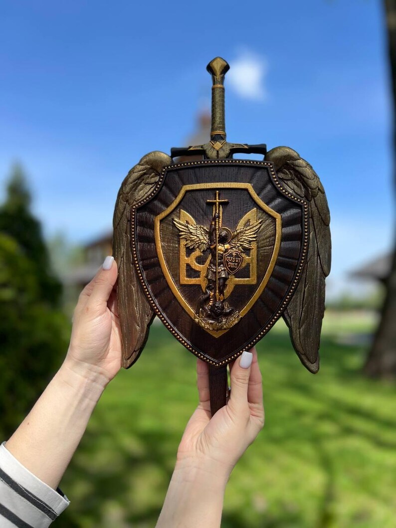 May include: A dark brown wooden shield featuring a central gold-toned relief of Saint Michael the Archangel and the Ukrainian trident symbol. The shield is adorned with large, textured gold wings on both sides and a sword handle at the top.