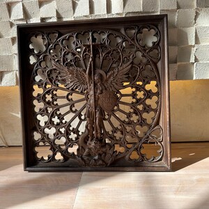 May include: A square dark brown wooden wall panel featuring an intricate openwork rose window design. At the center, a carved figure of an angel with wings holds a cross and a shield inscribed with the Latin text QUIS UT DEUS, standing over a figure.