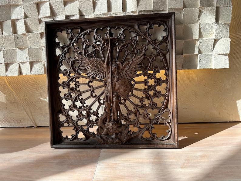 May include: A square dark brown wooden wall panel featuring an intricate openwork rose window design. At the center, a carved figure of an angel with wings holds a cross and a shield inscribed with the Latin text QUIS UT DEUS, standing over a figure.