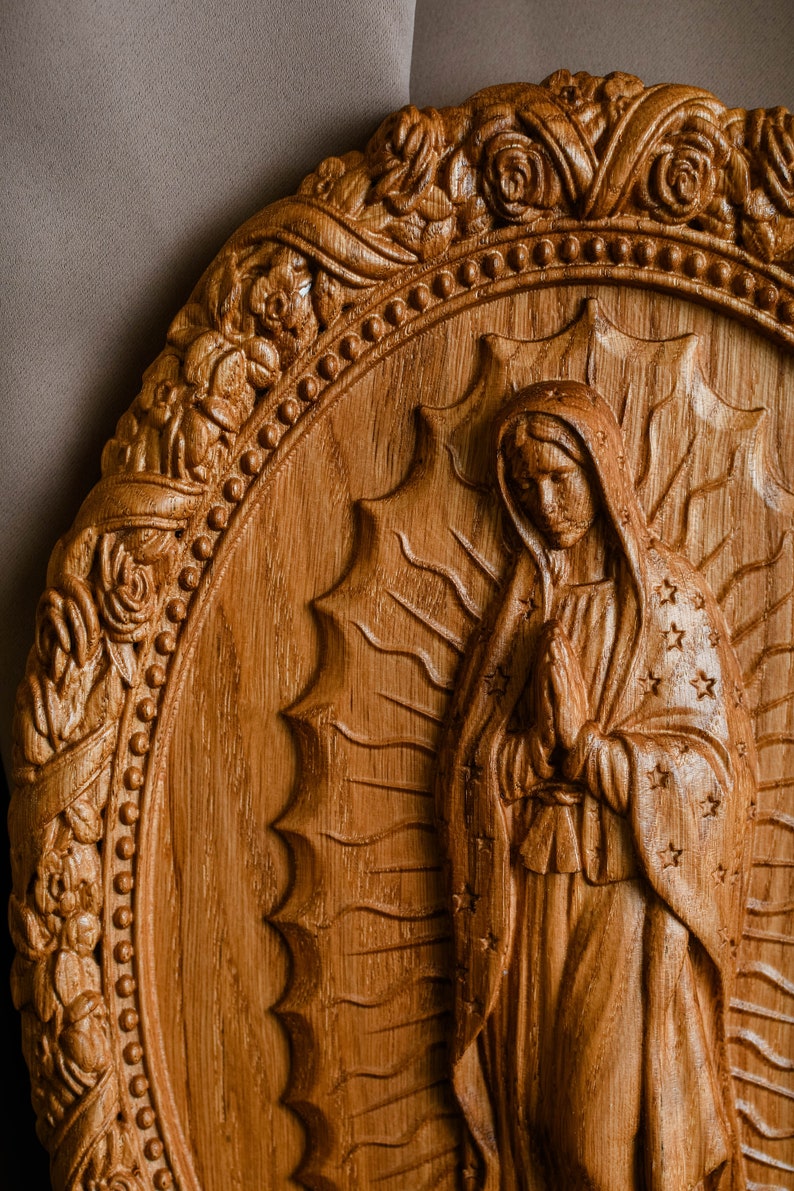 May include: A detailed wooden carving of a religious figure, featuring an oval frame with floral and beaded accents. The central figure is depicted with hands clasped in prayer, wearing a draped garment adorned with stars. The carving is in shades of brown.