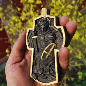 May include: A wooden cross with a carved depiction of an angel holding a sword, framed with gold trim. The angel is wearing armor and has a halo. The cross is held in a hand against a blurred background of yellow flowers and a red structure.
