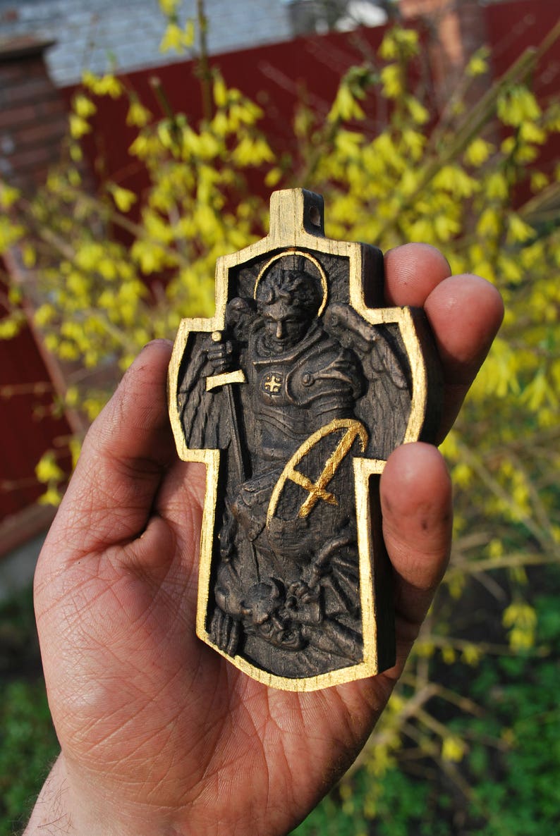 May include: A wooden cross with a carved depiction of an angel holding a sword, framed with gold trim. The angel is wearing armor and has a halo. The cross is held in a hand against a blurred background of yellow flowers and a red structure.