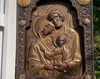 May include: A carved wooden icon depicting the Holy Family. The icon is framed in a dark brown wood with gold accents. The figures of Mary, Joseph, and Jesus are carved in relief and painted in gold and brown tones. The icon is a traditional religious artwork.