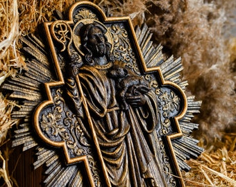May include: A wooden carving of Jesus holding a lamb, with a gold-painted background and a decorative frame. The carving is set against a backdrop of straw and dried grasses.