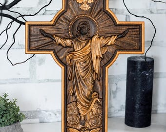 May include: A dark brown wooden cross featuring a central relief carving of a figure with arms outstretched, surrounded by a sunburst pattern. A golden dove is carved at the top, and a small winged cherub head is carved at the bottom of the cross.