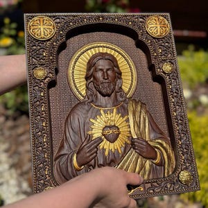 K&ouml;nnte beinhalten: Eine rechteckige Wandtafel aus Holz mit einem geschnitzten Relief des Heiligsten Herzens Jesu. Die Figur hat einen goldenen Heiligenschein und ein goldenes Herz, mit dunklen Holzt&ouml;nen und kunstvollen floralen Randverzierungen.