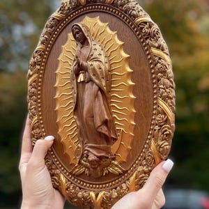 May include: A wooden oval plaque featuring a carved depiction of the Virgin Mary. The figure is in a brown robe with a gold halo and star accents. The frame is ornate with roses and gold accents.