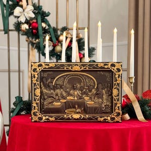 May include: A framed painting depicting the Last Supper with Jesus and his disciples. The painting is in a dark brown frame with gold accents. The painting is on a red tablecloth with white candles in front of it.