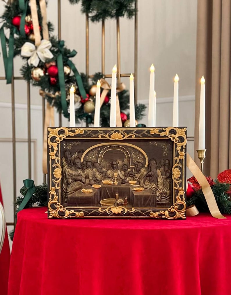 May include: A framed painting depicting the Last Supper with Jesus and his disciples. The painting is in a dark brown frame with gold accents. The painting is on a red tablecloth with white candles in front of it.