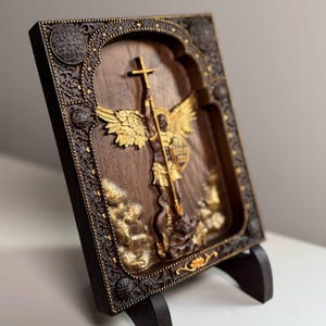May include: A wooden relief carving of an angel with gold-colored wings, holding a cross and spear. The artwork is set within a decorative frame with ornate detailing and is displayed on a wooden stand. The color palette includes shades of brown and gold.