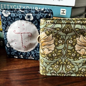 Peut inclure: Deux pochettes recouvertes de tissu avec un motif floral. La pochette de gauche est bleue avec des fleurs blanches et un cercle blanc avec une lettre T brodée en rouge. La pochette de droite est verte avec un motif floral dans des tons de vert, marron et blanc.