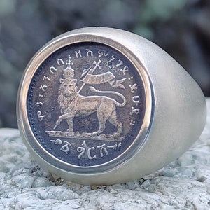 Może przedstawiać: Srebrny pierścionek z okrągłym, wypukłym wzorem przedstawiającym lwa z koroną i sztandarem. Wzór otoczony jest tekstem w obcym języku.