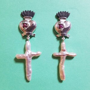 May include: A pair of silver earrings with a heart and cross design. The heart is topped with a black crown and features a red gemstone. The cross is made of a white pearl.