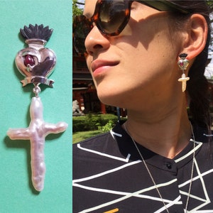 May include: A pair of silver earrings featuring a heart and cross design. The heart is black and silver with a red gemstone. The cross is made from a white pearl-like material.
