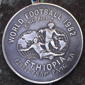 Puede incluir: Una moneda conmemorativa redonda de color plateado que muestra a dos futbolistas en acción sobre un globo terráqueo. El texto dice WORLD FOOTBALL 1982, Birr 2, ETHIOPIA e incluye escritura amhárica. El diseño está grabado en metal.