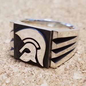 Puede incluir: Un anillo de plata con una imagen estilizada en relieve de un casco en la parte delantera. El anillo tiene una banda cuadrada con cuatro líneas en relieve en el lateral.